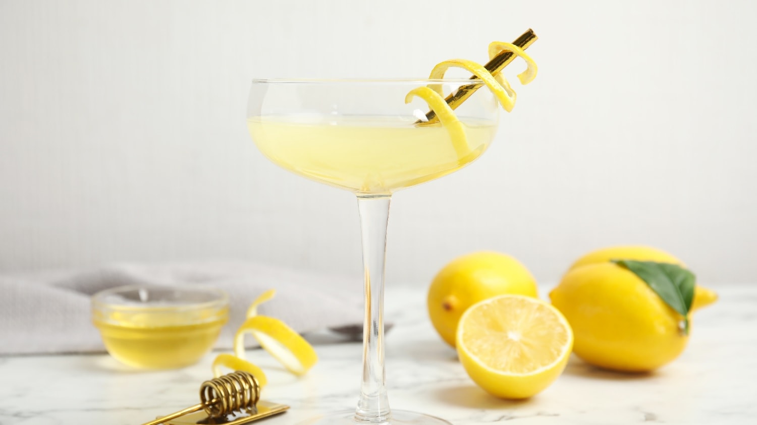 Ein Coupette-Cocktailglas mit dem gelben Drink und einer Zitronenschale. Am hellen Boden sind halbierte Zitronen und ein kleines Glas mit Honig.