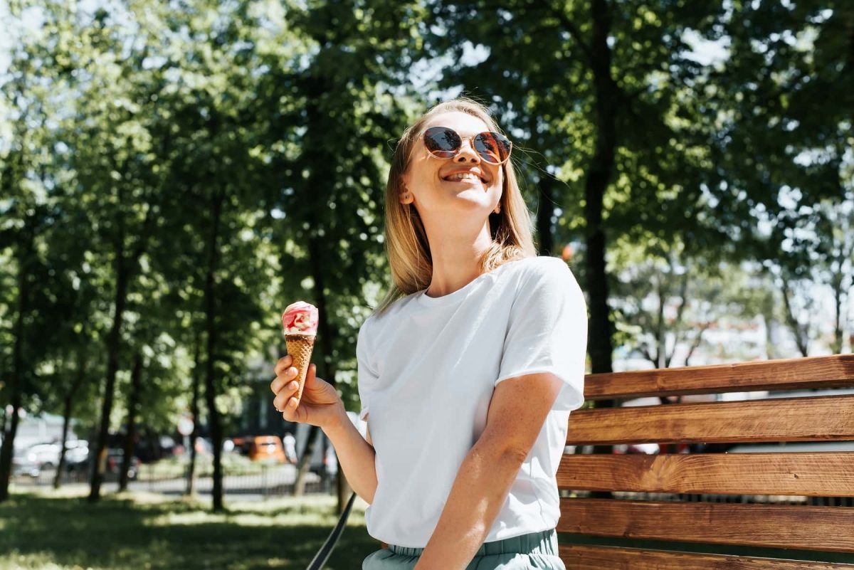 Eisdielen in Berlin: Eine lachende Frau sitzt im Sommer auf einer Parkbank und hält ein Eis in der Hand.