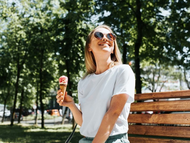 Eisdielen in Berlin: Eine lachende Frau sitzt im Sommer auf einer Parkbank und hält ein Eis in der Hand.