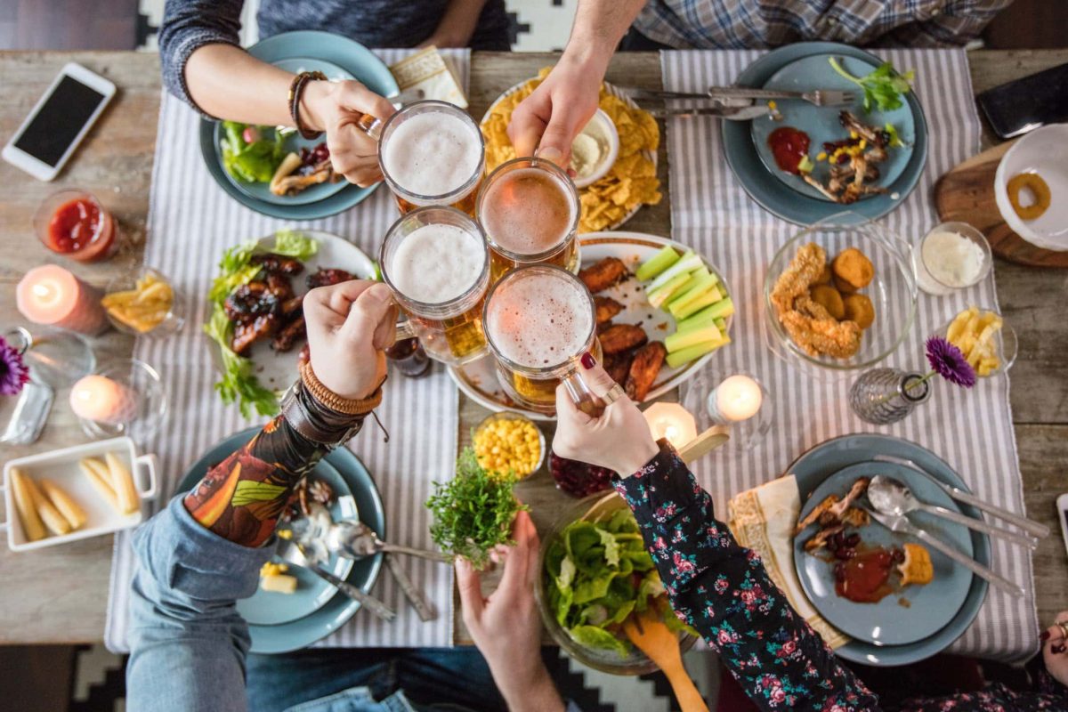 Wieviel Fleisch pro Person soll man kaufen? Freunde stoßen über gedecktem Tisch mit diversem Speisen.