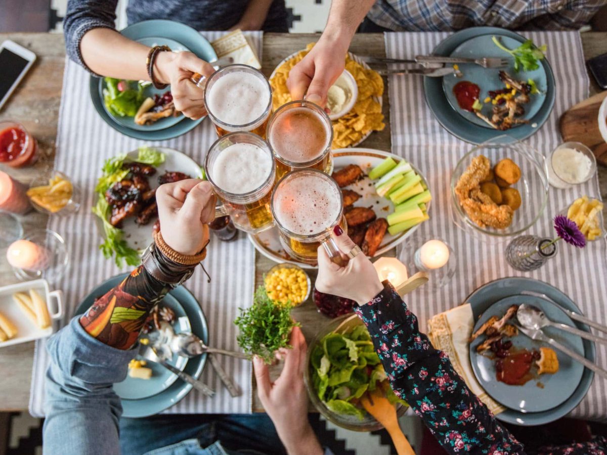 Wieviel Fleisch pro Person soll man kaufen? Freunde stoßen über gedecktem Tisch mit diversem Speisen.
