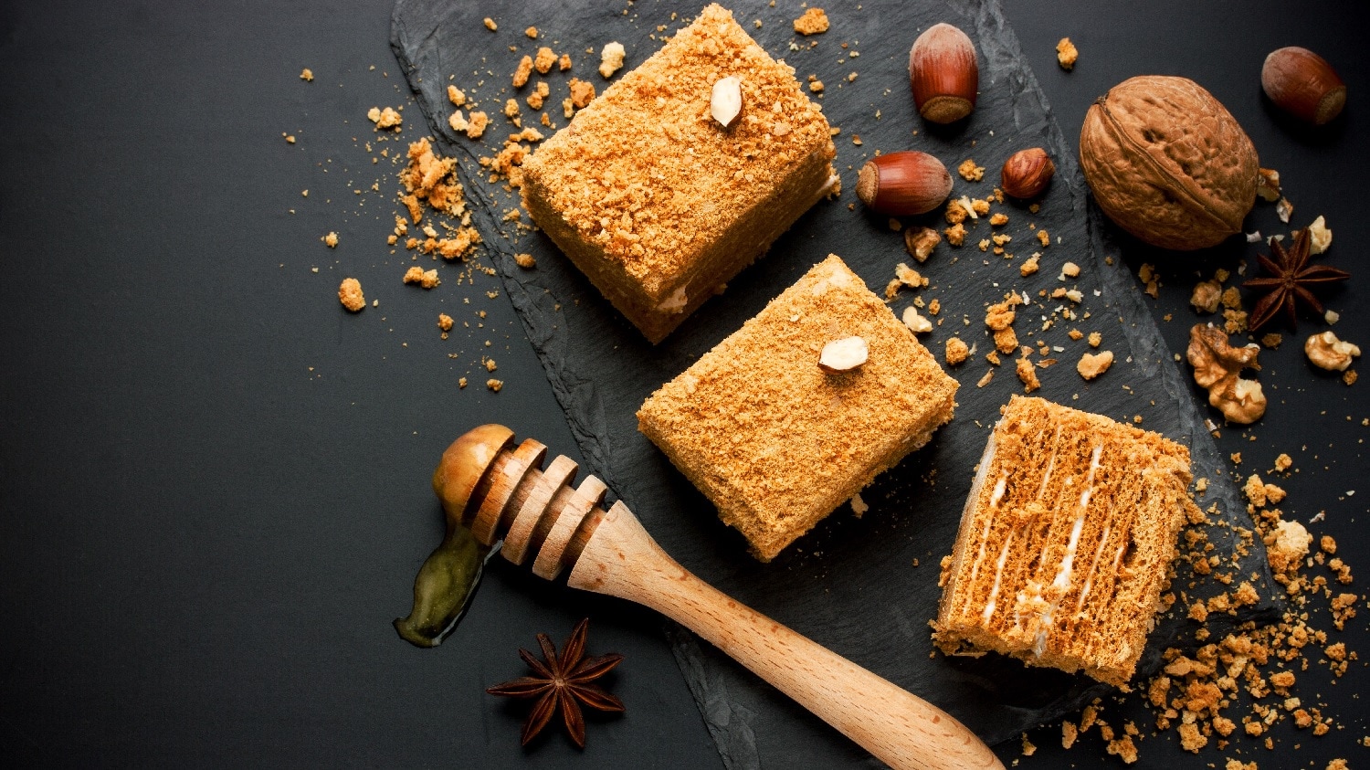 Drei Stücke des Honigkuchens auf einer dunklen Schieferplatte. Darum verteilt sind Nüsse und ein Holzstab mit Honig.