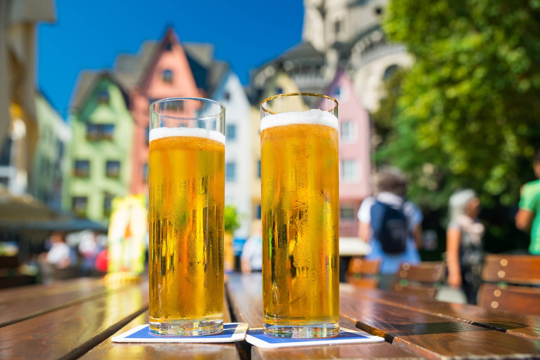 Kölner Bierbörse: Zwei Kölsch auf einem Biergartentisch, im Hintergrund unscharf die bunten Häuserfassaden am Rheinufer.