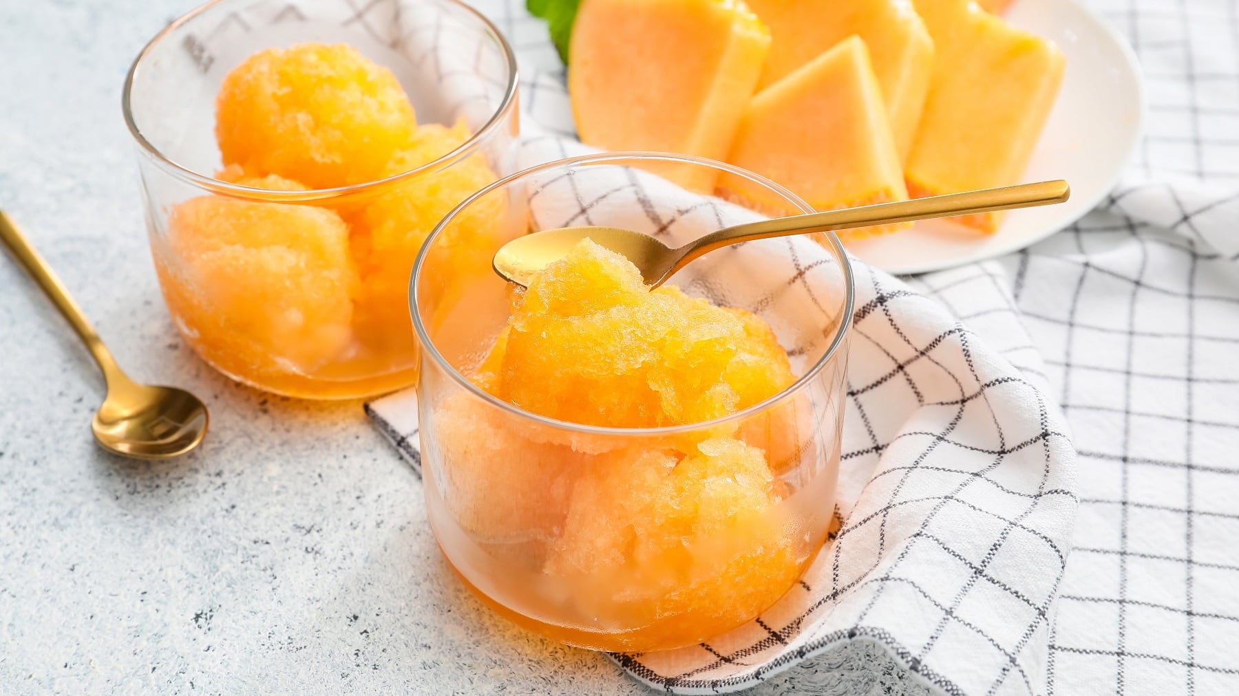 Zwei Gläser Melonensorbet mit Mango und zwei goldenen Teelöffeln, daneben ein Teller Melonenstücke und ein Geschirrtuch.