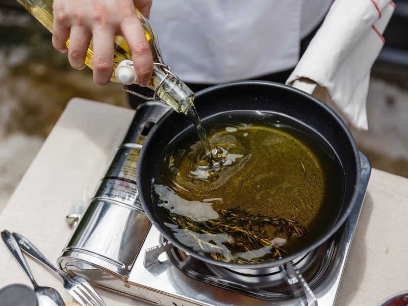 Ein Koch gibt Olivenöl in eine Pfanne. In dem Olivenöl liegen Rosmarin-Zweige. Die Pfanne steht auf einer Gasplatte.