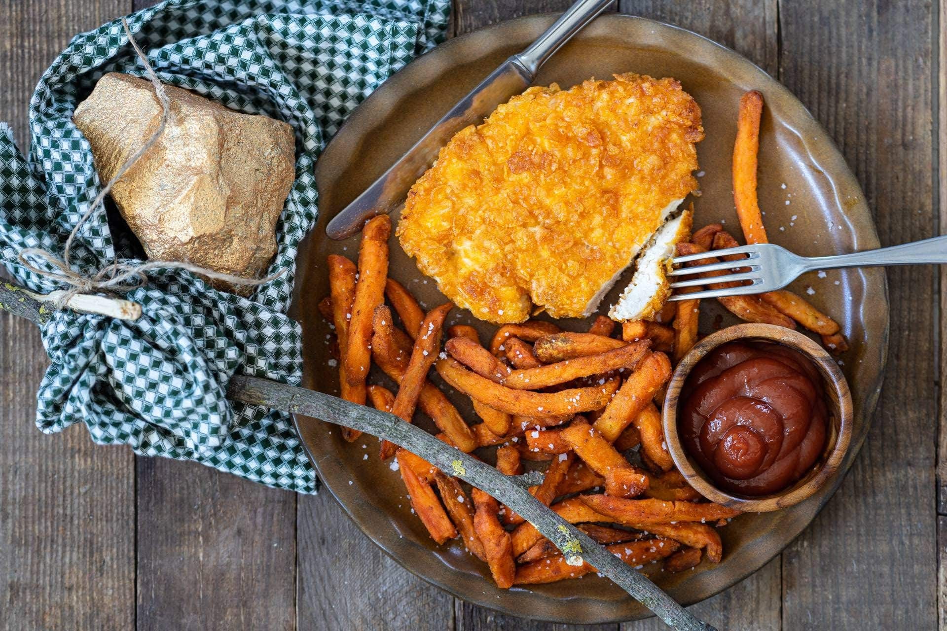 Ein Schnitzel mit Cornflakes-Panade, Süßkartoffeln und einer Schale Ketchup auf einem Teller in der Draufsicht, daneben ein Tuch mit einem kleinen Goldklumpen.
