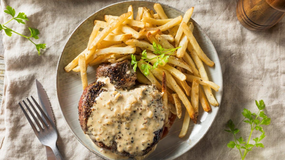 Ein heller Teller mit Pommes und dem Steak au poivre auf einem beigen Tuch. Dazu ein paar stängel Petersilie die auf dem Tisch verteilt sind.