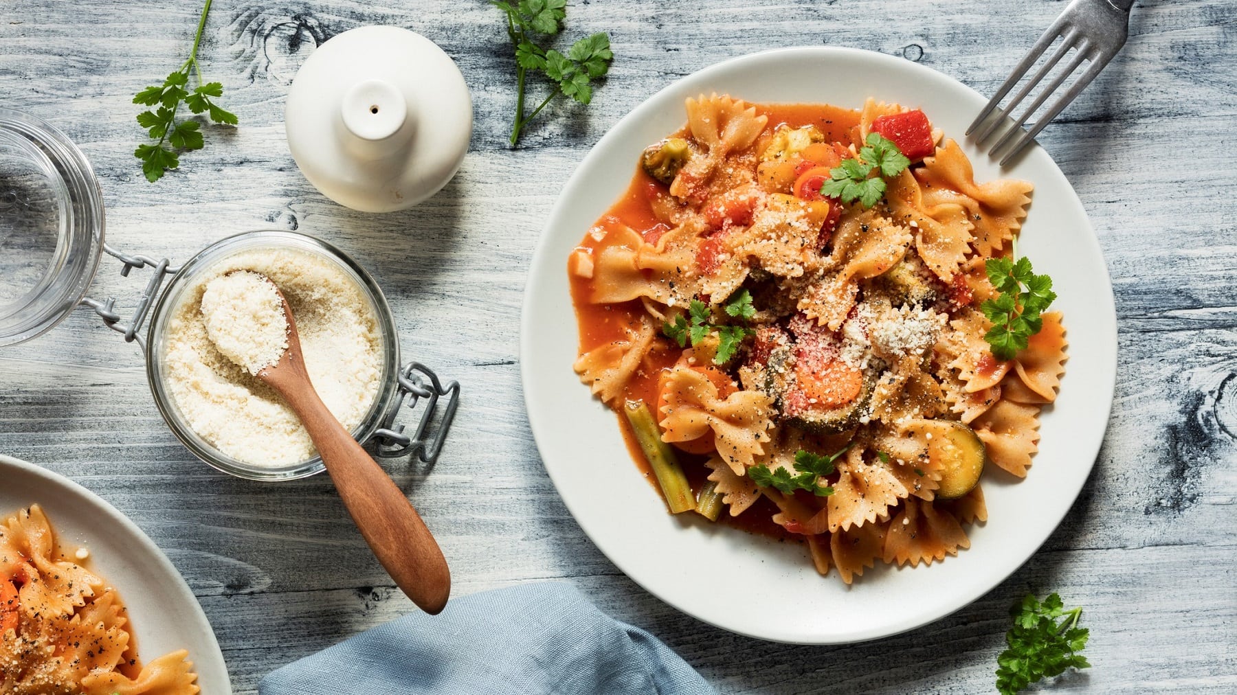 Ein Teller vegane Pasta mit Gemüseragout in der Draufsicht, daneben eine Gabel und ein Glas mit Parmesan, außerdem frische Kräuter und ein Salzstreuer.