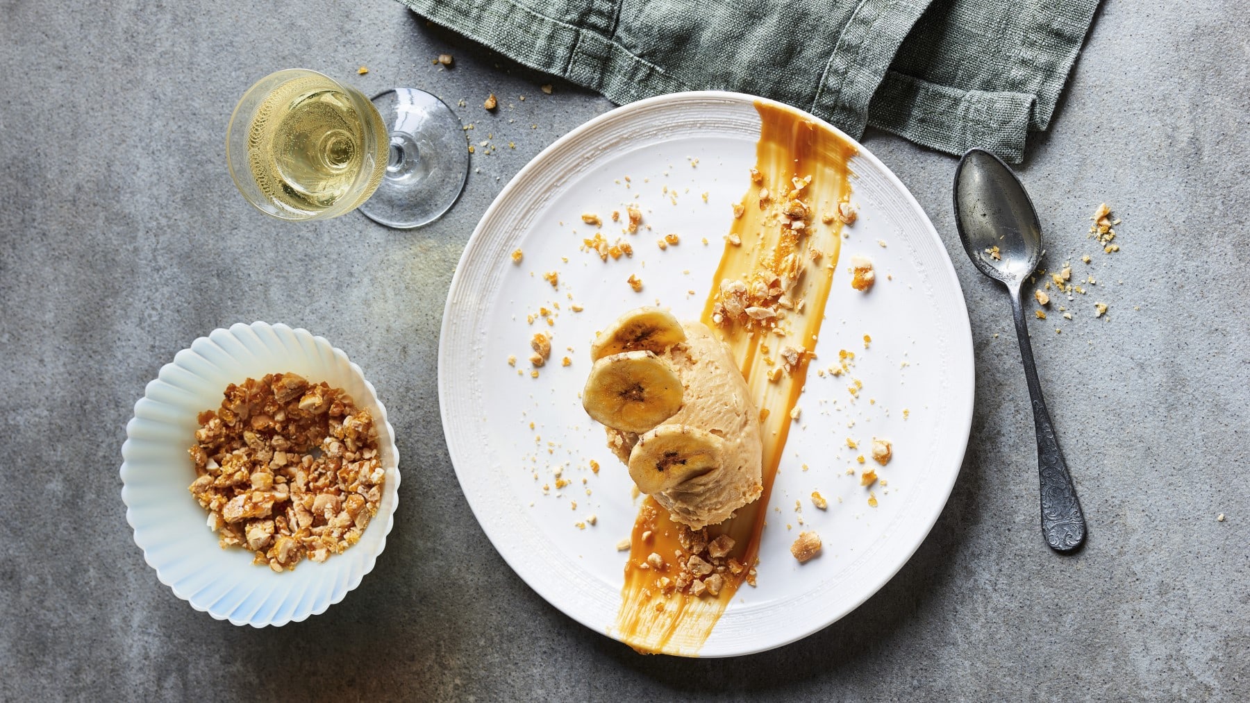 Erdnussbutterparfait mit Salted-Caramel-Crunch, serviert auf einem weißen Teller mit Bananen garniert und mit Wein auf einem Steintisch serviert.
