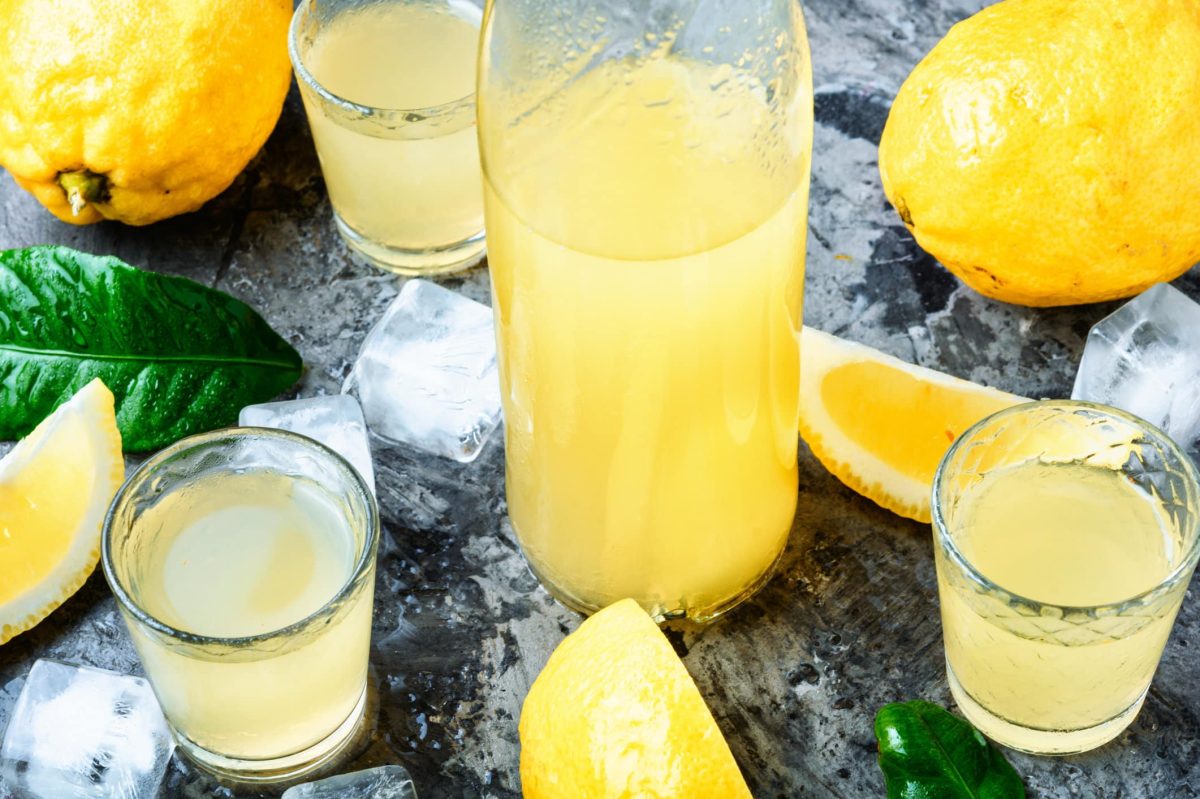Limoncello in Gläsern und Flaschen auf dunklem Untergrund. Drum herum liegen Zitronen und Eiswürfel.