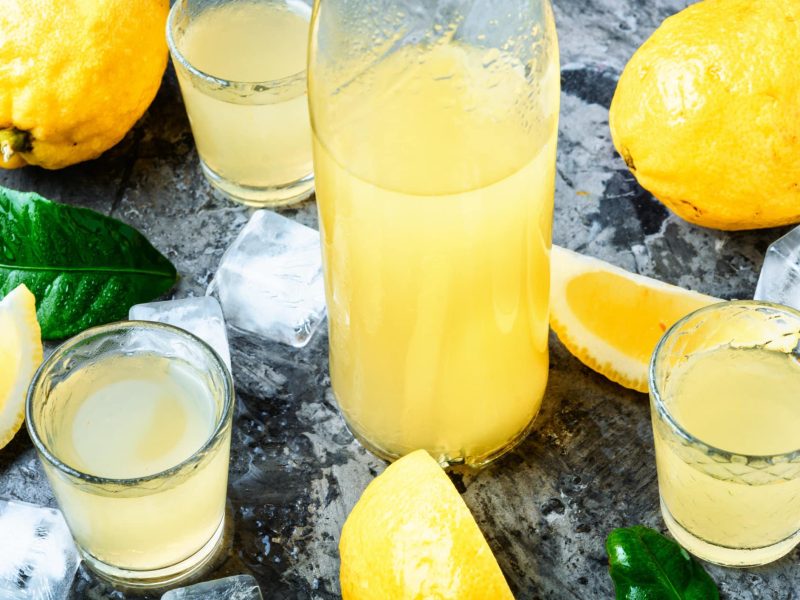 Limoncello in Gläsern und Flaschen auf dunklem Untergrund. Drum herum liegen Zitronen und Eiswürfel.