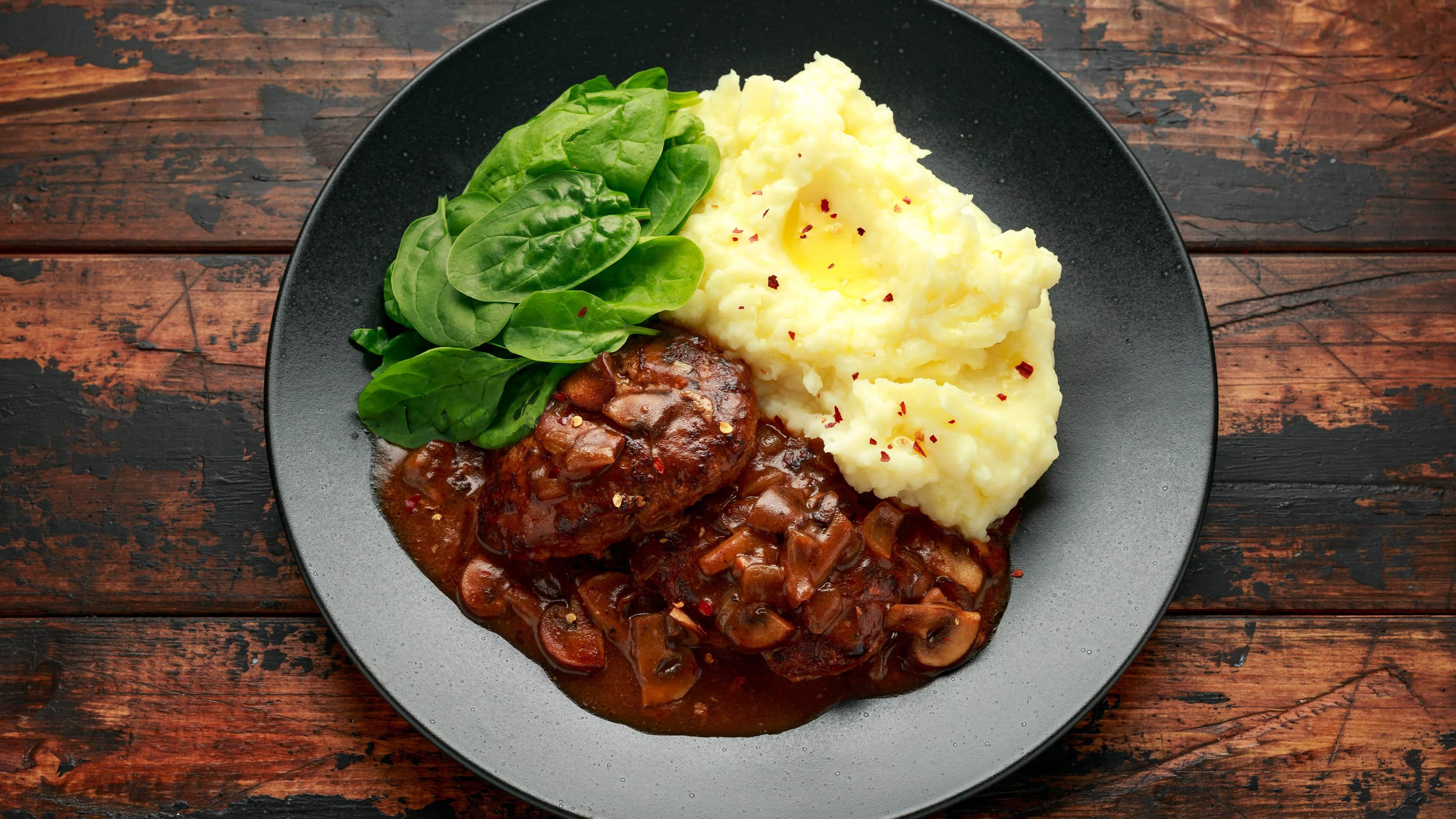 Salisbury Steak mit Kartoffelbrei auf schwarzem Teller. Dieser steht auf Holztisch.