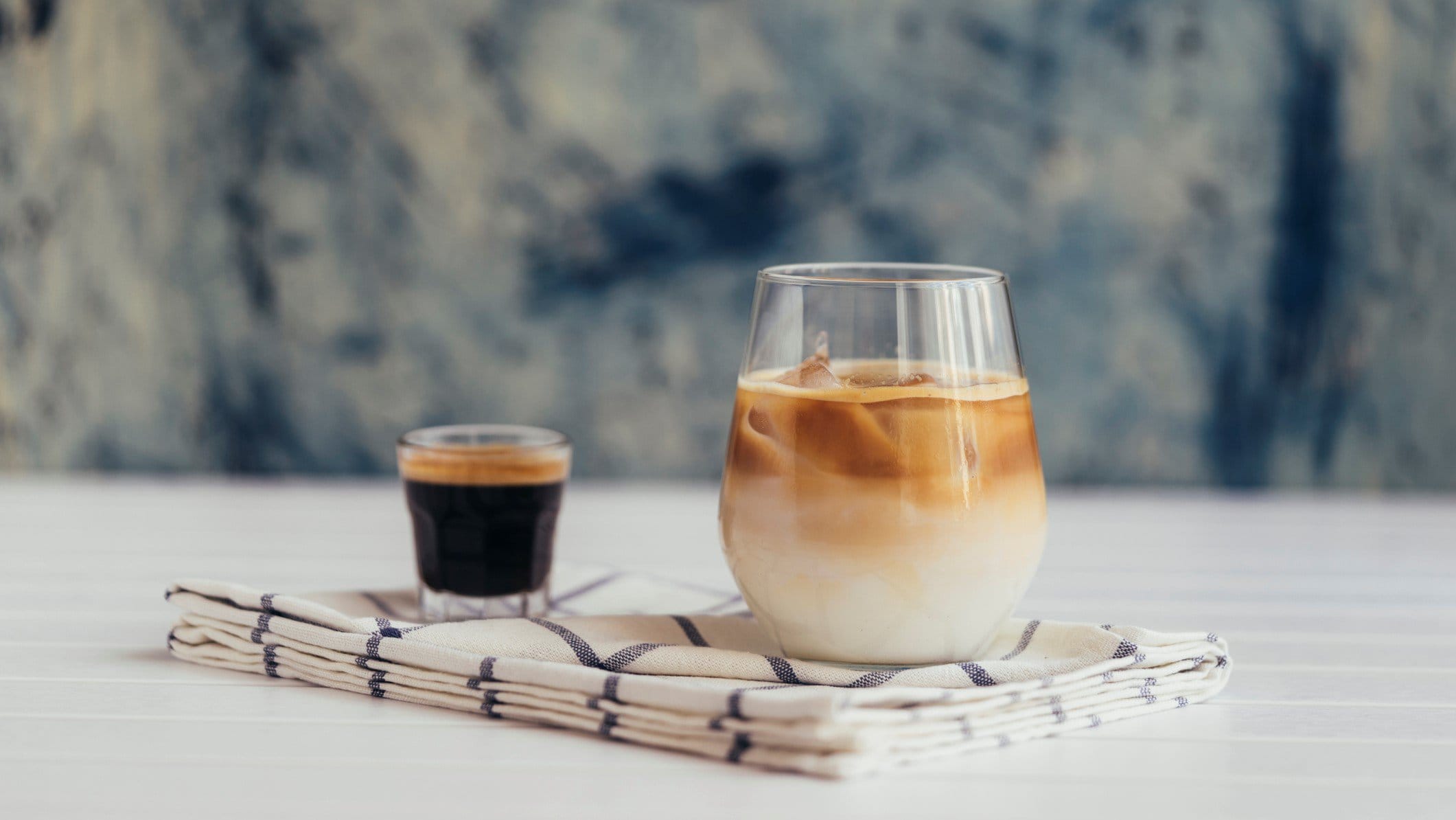 Cold Johnny Eiskaffee in einem Glas mit Espresso auf Tuch und hellem Tisch vor blau-weißem Hintergrund. Frontalansicht.