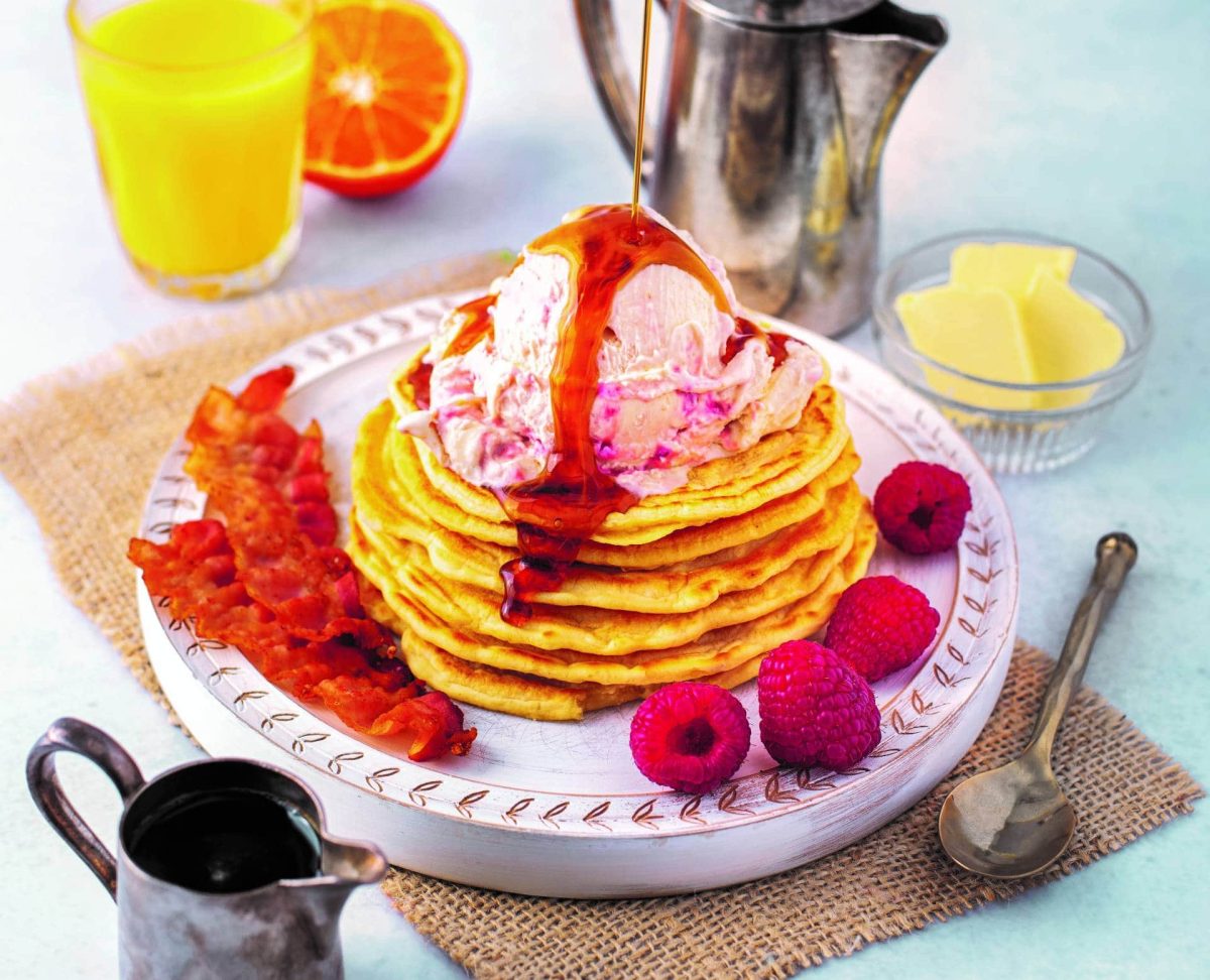 Ein Pancake-Turm mit American Breakfast Eis aus dem Kochbuch "Eis aus dem Wunderland", daneben frische Himbeeren und gebratener Speck, außerdem ein Glas O-Saft, Kännchen und ein Löffel.