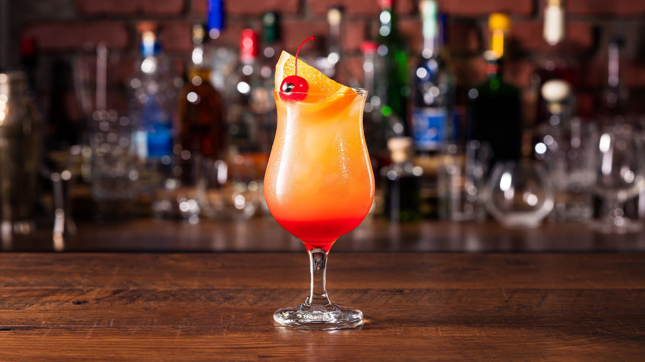 Tropischer Hurricane-Cocktail mit Rum und Maracuja in Tiki-Glas auf Theke. Im Hintergrund eine Bar mit Spirituosen. Frontalansicht.