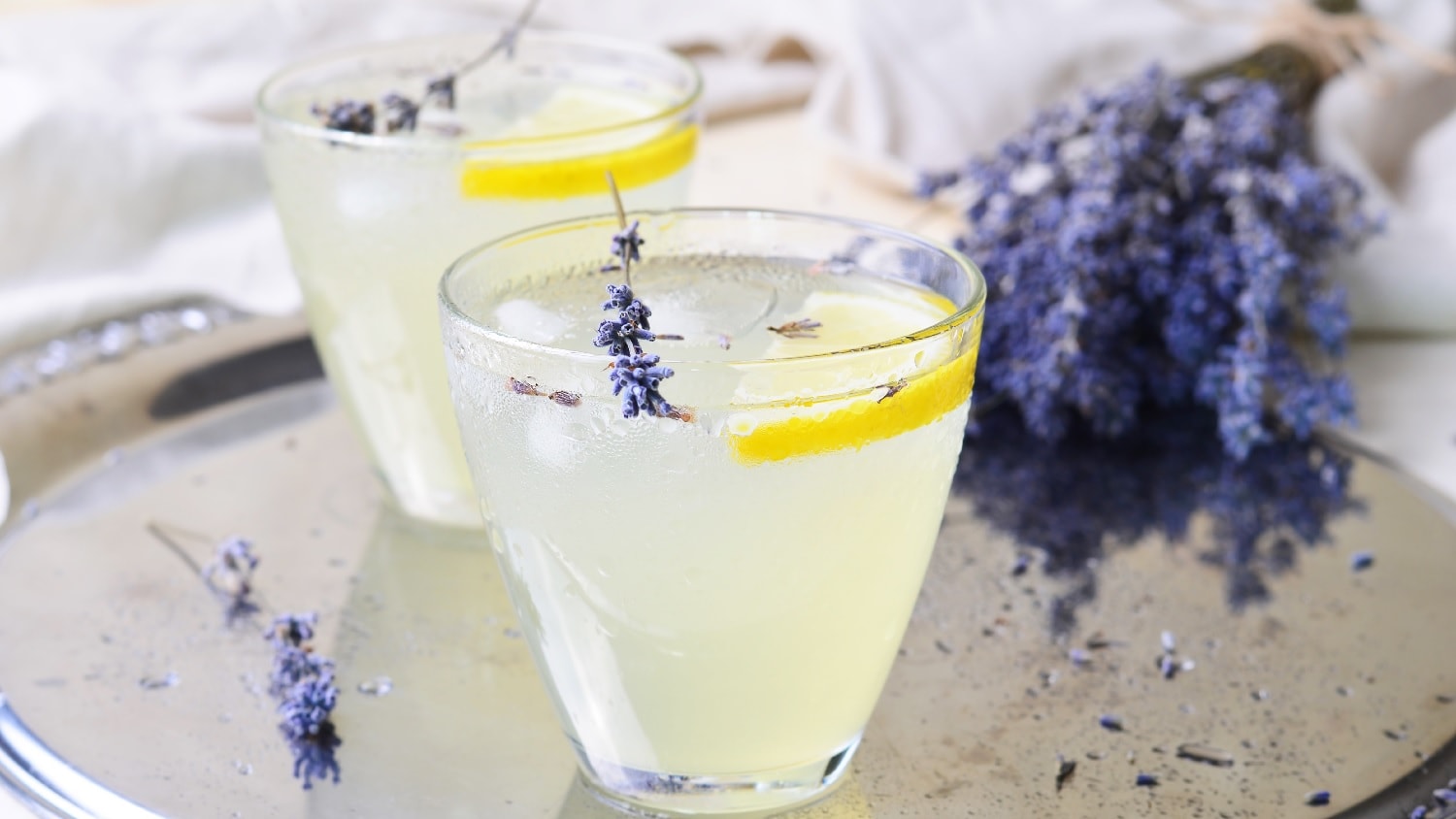 Zwei Gläser der Limonade mit Lavendelblüten auf einem Silbertablett und hellem Hintergrund. Ebenfalls im Hintegrund ist ein ganzer Strauß Lavendel.