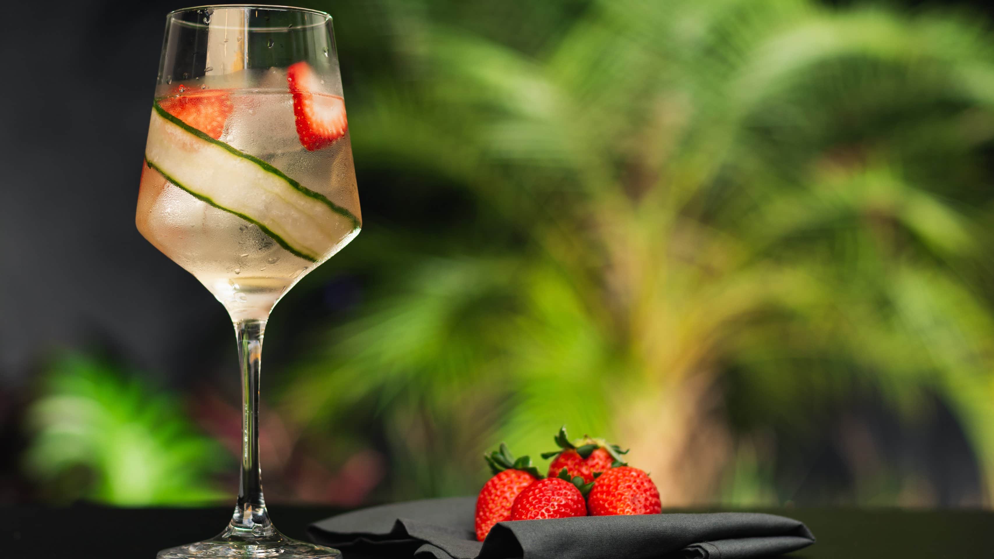 Lillet Gin Tonic in Weinglas auf dunklem Tisch vor unscharfem Hintergrund mit Pflanze. Daneben Erdbeeren. Frontalansicht.