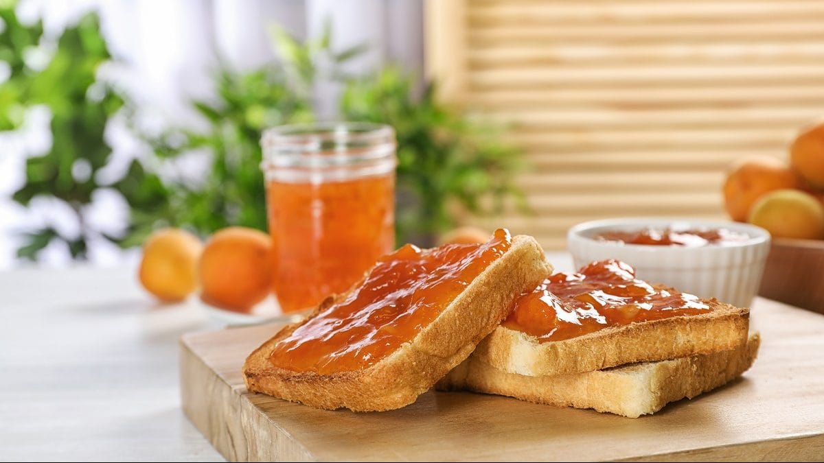 Ein Glas Marillen-Mohn-Marmelade steht auf einem Tisch. Daneben liegen zwei Toasts mit dem Aufstrich. Und auch Aprikosen liegen auf dem Tisch.