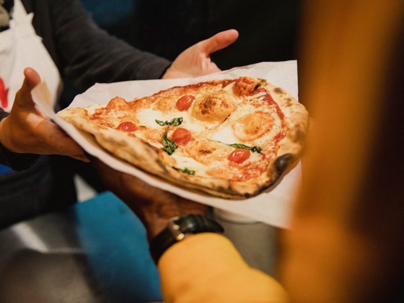 Beim True Italian Pizza Street Festival Person reicht Pizza auf Krepp an eine andere Person weiter. Frontalaufnahme.