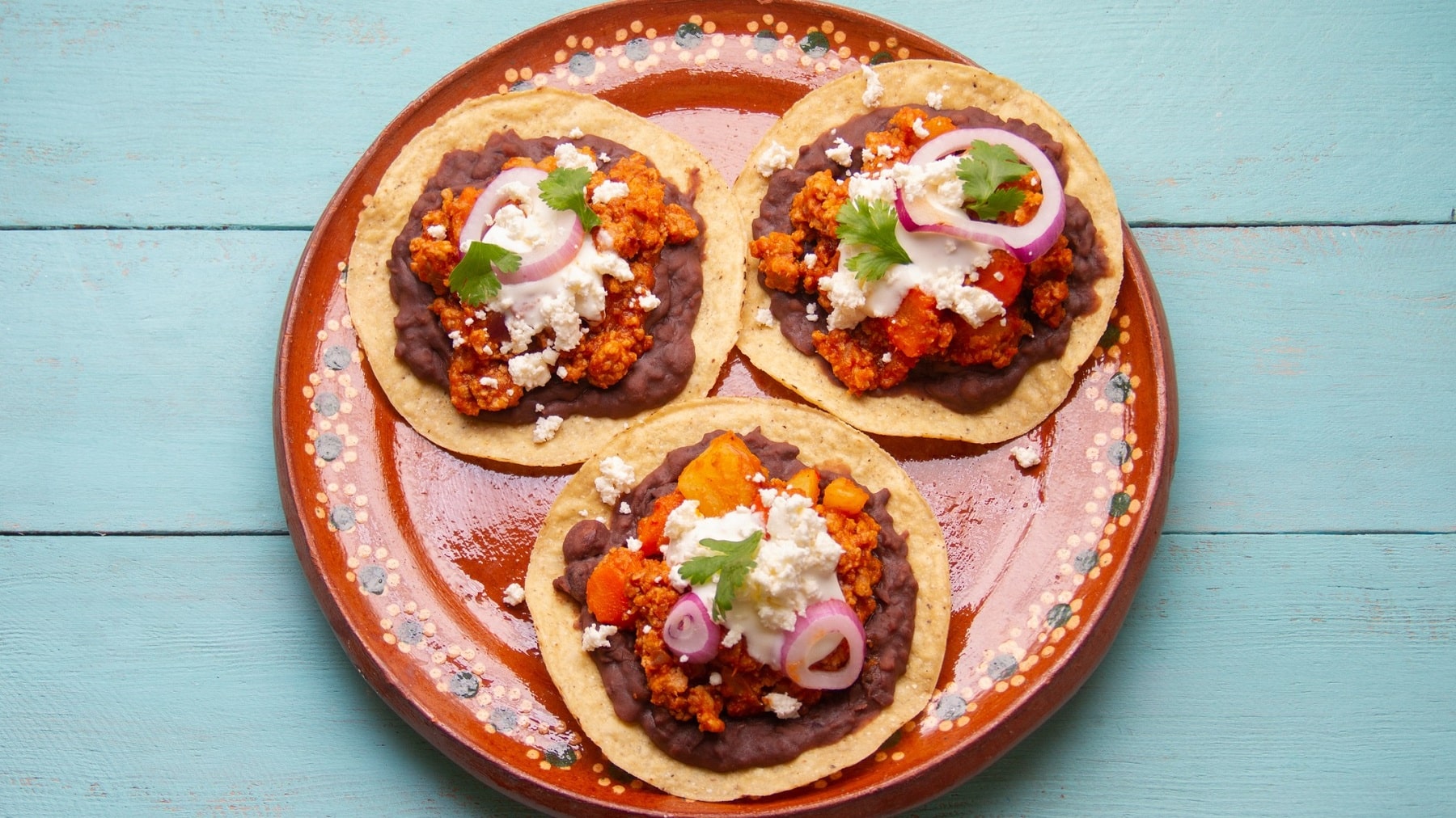 Drei vegane Hackfleisch-Tacos mit Bohnenmus, roten Zwiebeln, Koriander, saurer Sahne und Feta auf einem bunten Teller in der Draufsicht.