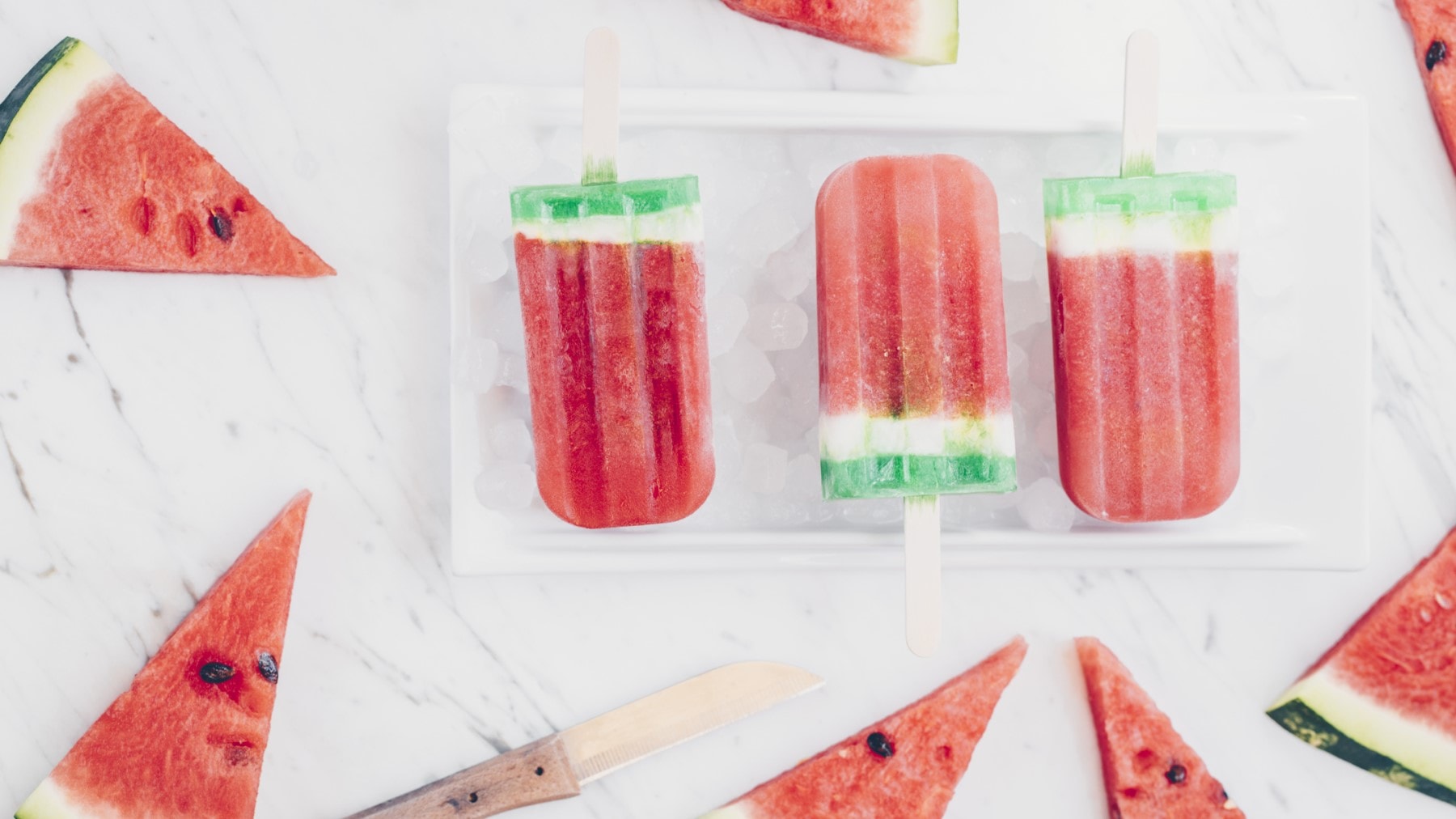 Wassermelonen Popsicles in einer Schale mit Eiswürfeln. Daneben liegen Wassermelonenscheiben.