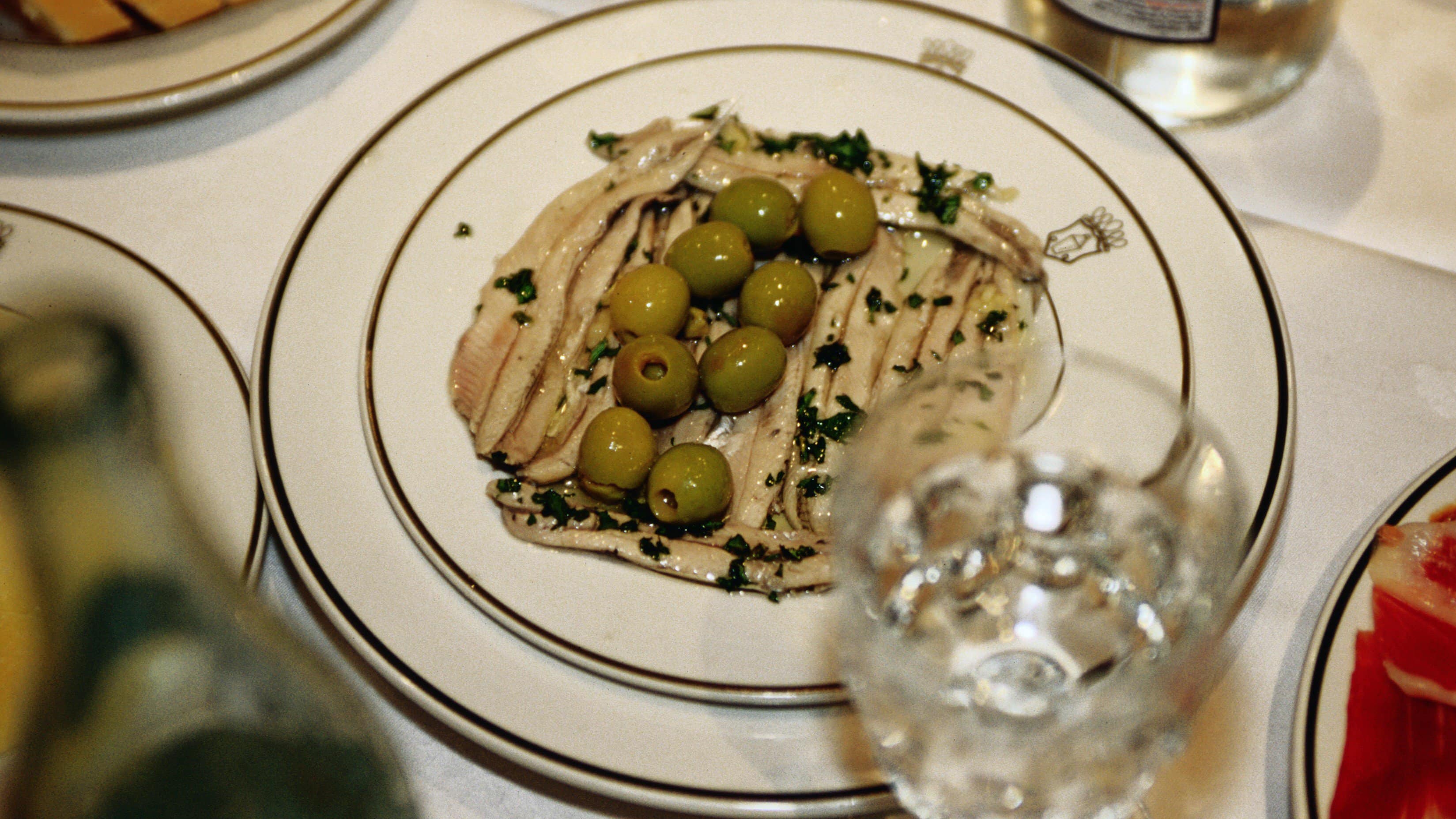 Boquerones en Vinagre eingelegte Sardellen auf weißem Teller und weißem Tisch. Daneben ein Glas Wasser und weitere Tapas am Bildrand. Draufsicht.