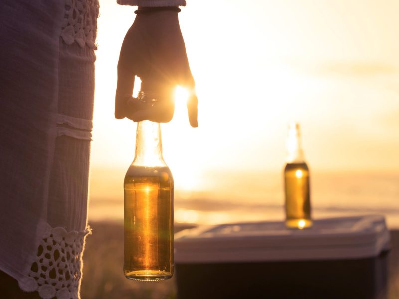 Frau hält Bier im Sonnenuntergang. Vor ihr steht eine weitere Flasche!