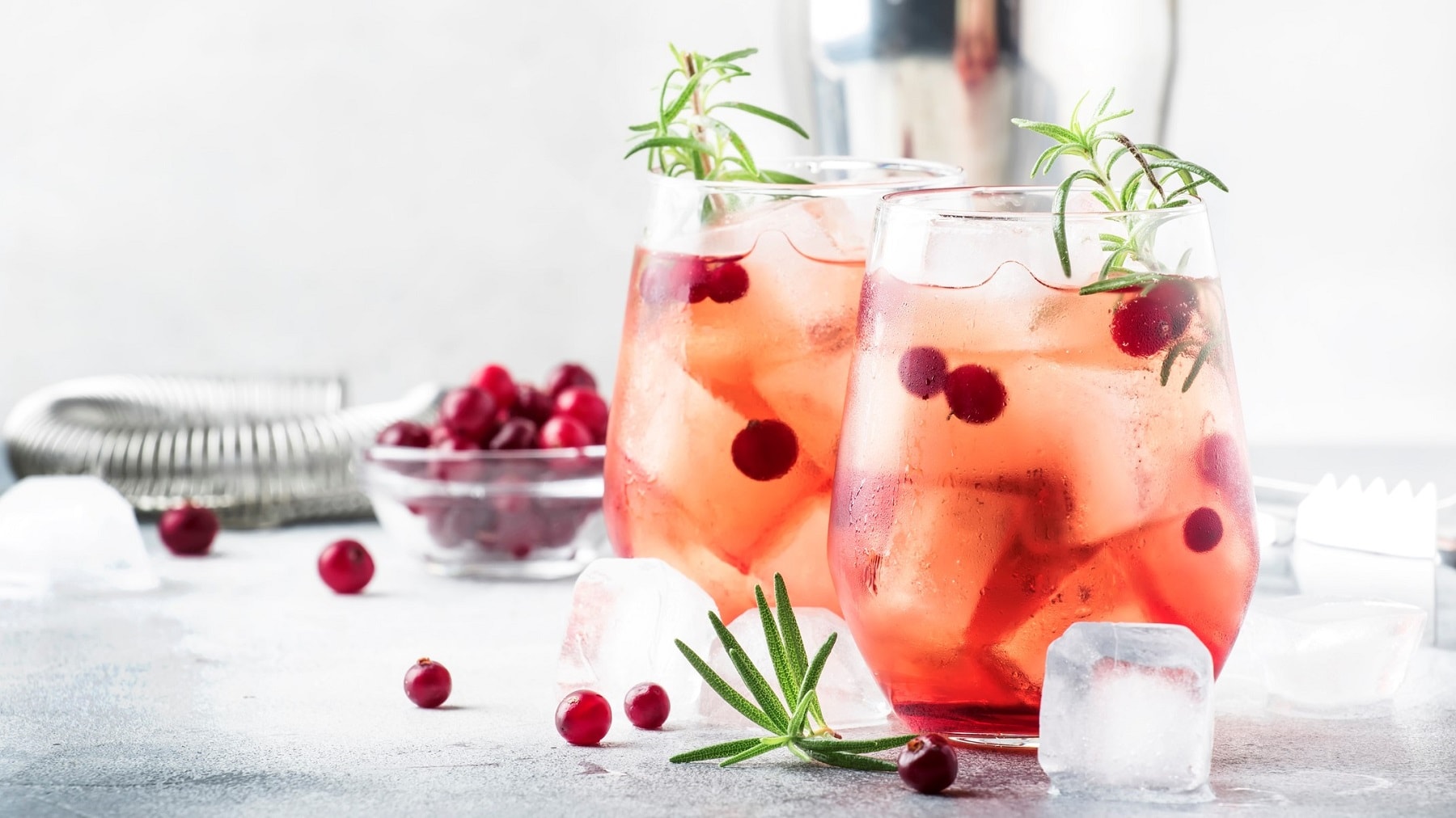 Zwei Gläser Berry-Lime-Cocktail mit Johannisbeeren, Eis und je einem Zweig Rosmarin. Drumherum Eiswürfel und Bar-Equipment.