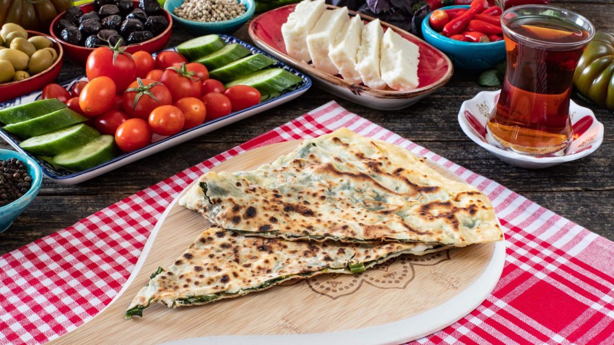 Gözleme auf Holzbrett auf rotem Tuch auf mit türkischen Speisen gedecktem Tisch. Daneben Tee. Draufsicht.