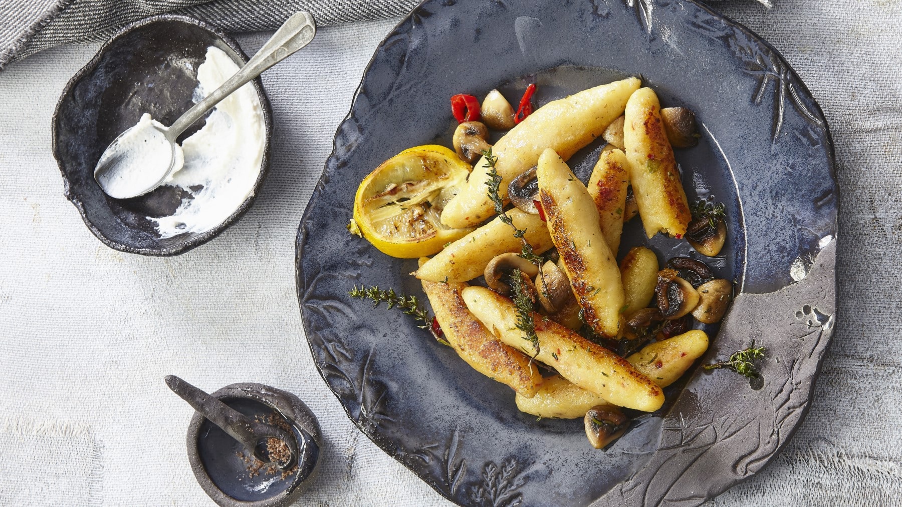 Kartoffelgnocchi mit Champignons und Sauerrahm garniert mit Zitrone, Thymian und Chili auf einem dunklen Teller neben einer Schale mit Sauerrahm vor grauem Hintergrund.
