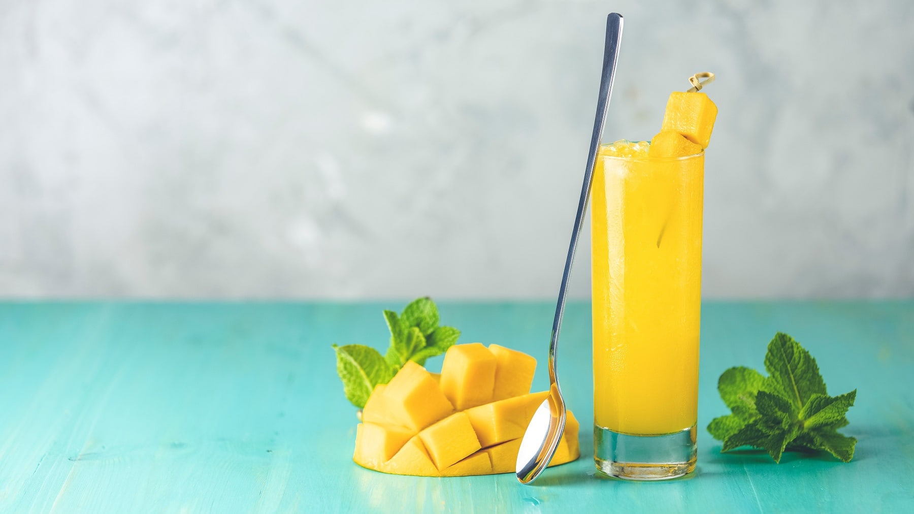 Ein Glas Mango-Limonade mit Ginger Beer und frischen Mangostücken, daneben liegen Minzblätter und eine aufgeschnittene Mango.