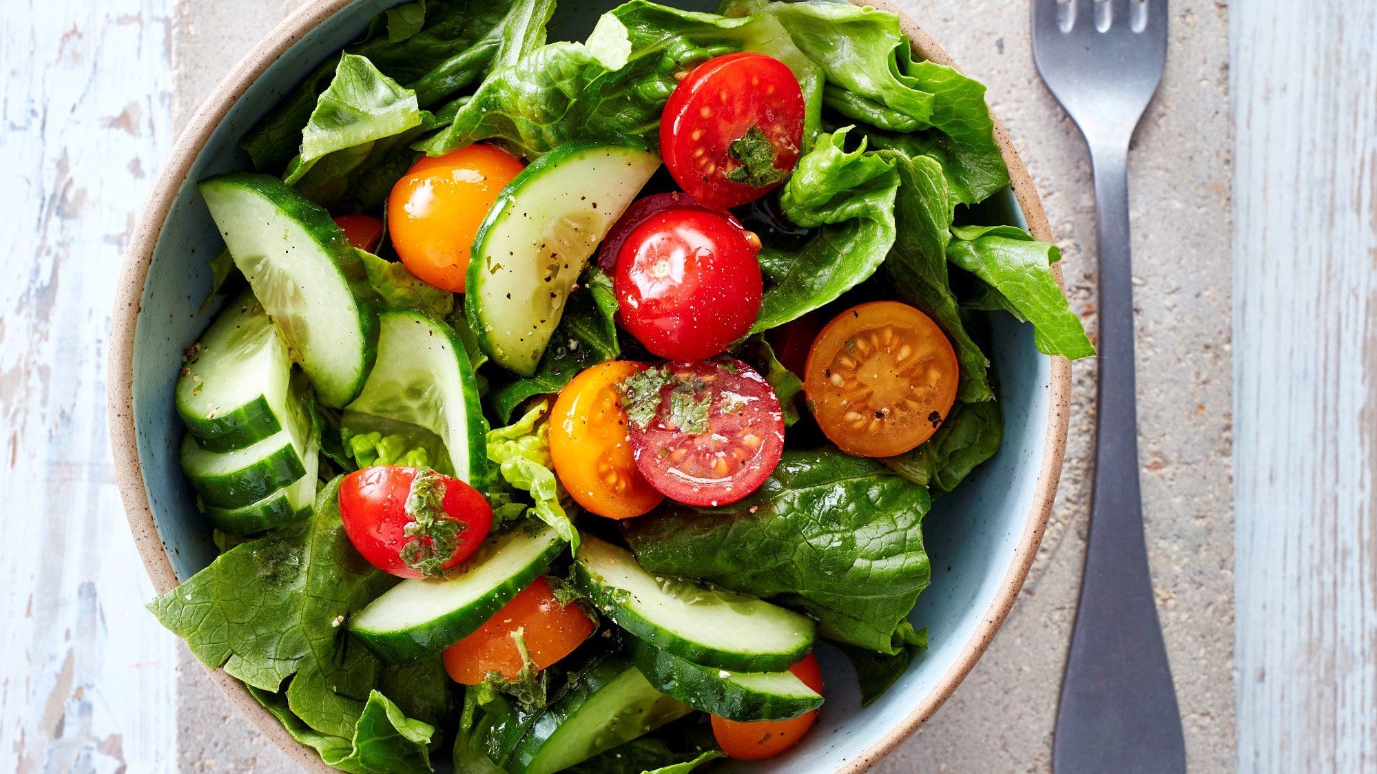 Schüssel mit grünem Salat, Tomaten und Gurkenscheiben und Minzdressing neben einer Gabel vor hellem Hintergrund.