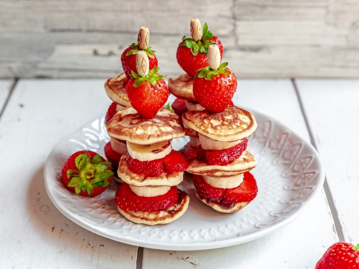 Pancake-Kebabs, auf Spieße aufgesteckte kleine Pancakes mit Erdbeer- und Bananenscheiben, auf einem Teller angeordnet.