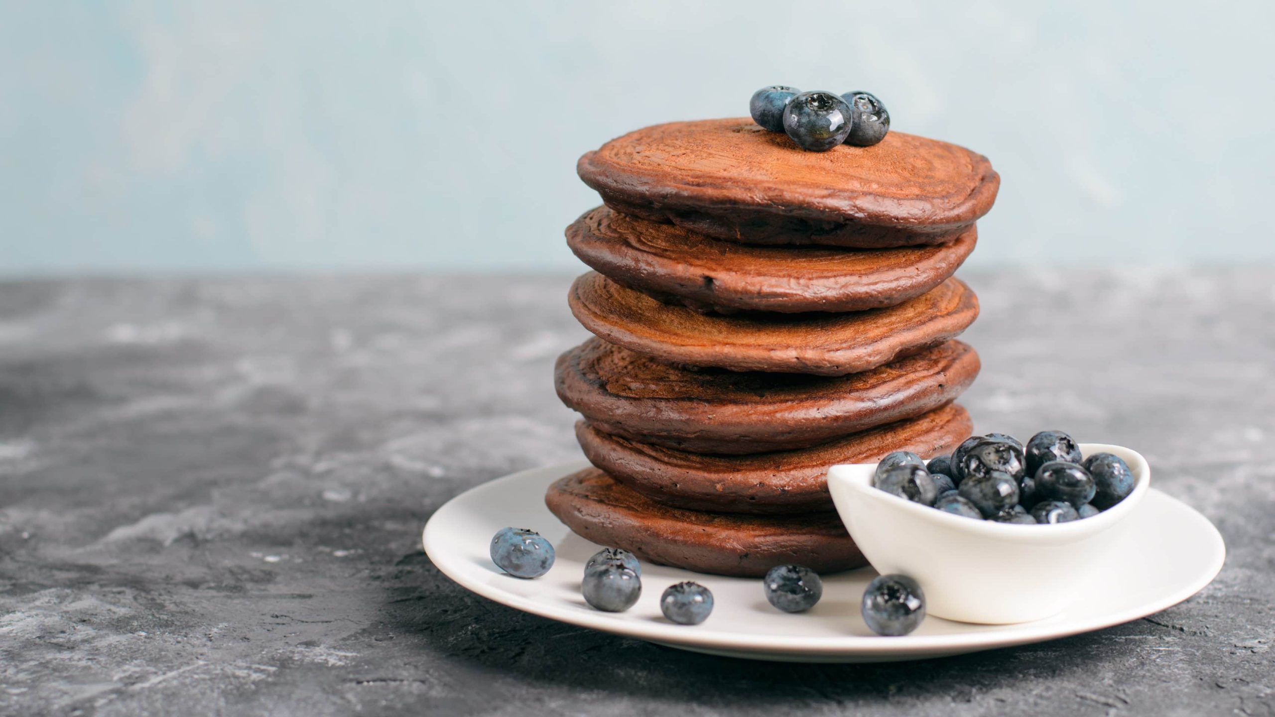 Ein weißer Teller mit den Schokoladen-Pancakes und Blaubeeren auf einem grauen Untergrund.