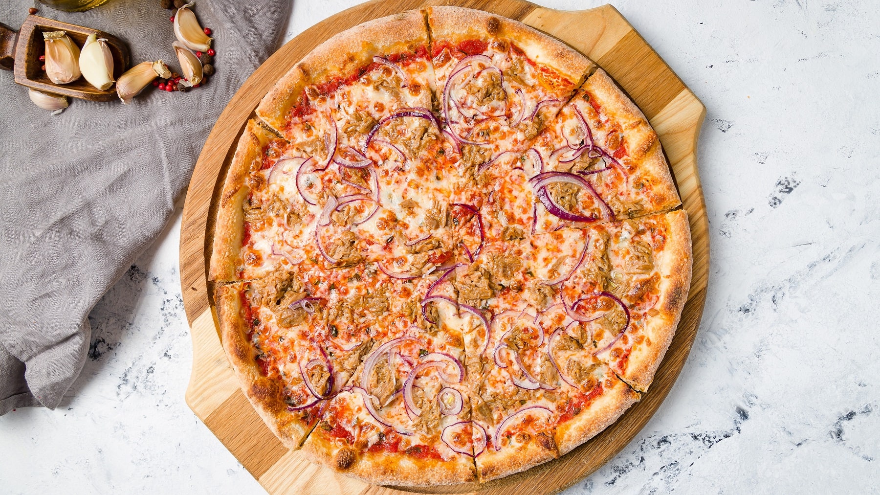 Eine geschnittene vegane Thunfischpizza mit roten Zwiebeln auf einem runden Holzbrett in der Draufsicht, daneben lieben Knoblauchzehen und ein graues Geschirrtuch.