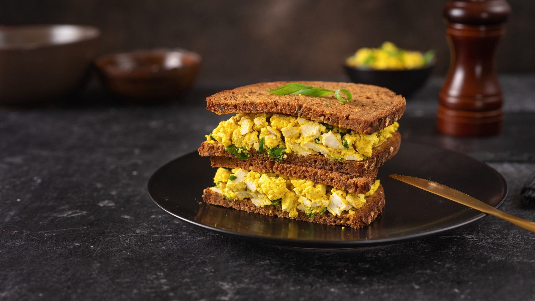 Zwei Sandwiches mit dunklem Brot und veganem Eiersalat gestapelt auf einem Teller, daneben liegt ein goldenes Messer.