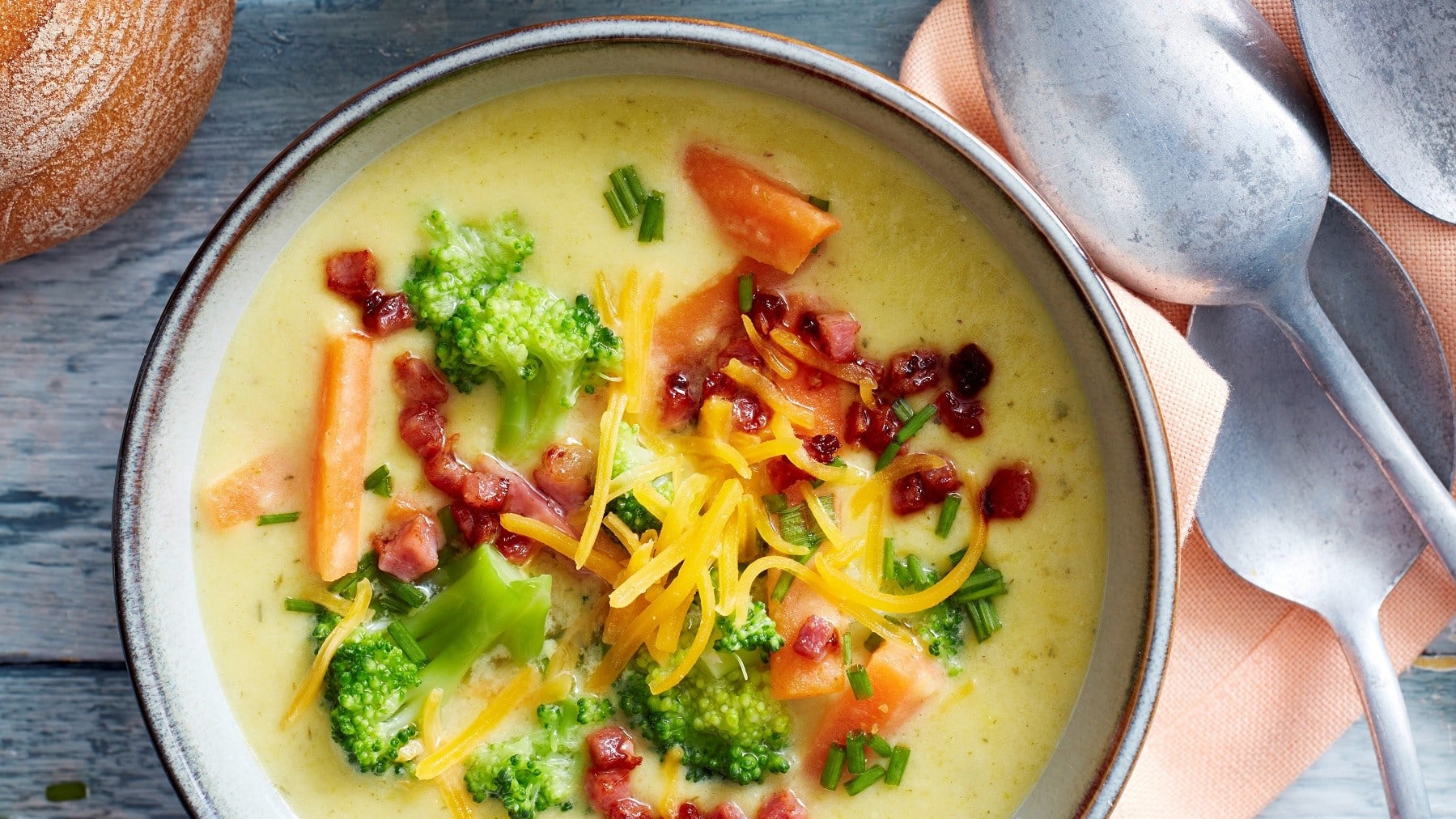 Cremige Cheddar-Brokkoli-Suppe, garniert mit Speck, frischem Brokkoli und Schnittlauch, serviert in einer Tonschale auf einem grauen Holztisch.