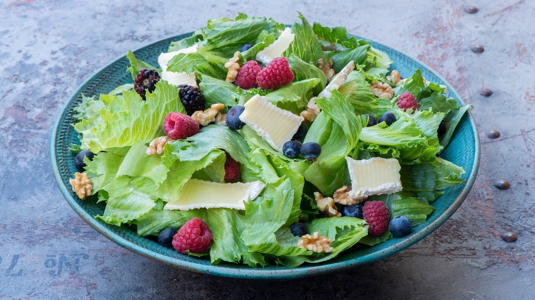 Endivien-Salat mit Beeren und Brie, garniert mit Walnüssen und saurer Vinaigrette und serviert in einer großen, blauen Tonschale auf einem hellgrauen Schiefertisch