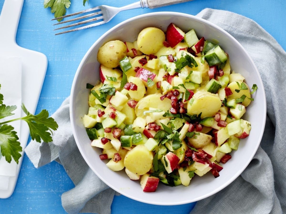Kartoffel-Gurken-Salat mit Apfel, Speckwürfeln und frischer Petersilie, serviert in einer großen Salatschale auf einem blauen Küchentisch.