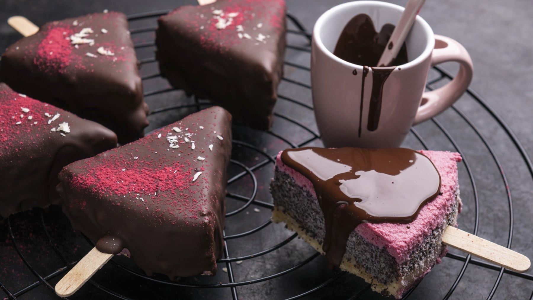 Ein paar vegane Cheesecake-Sticks mit Mohn und Himbeere überzogen mit Schokolade neben einer Tasse mit geschmolzener Schokolade auf einem Gitter vor dunklem Hintergrund.