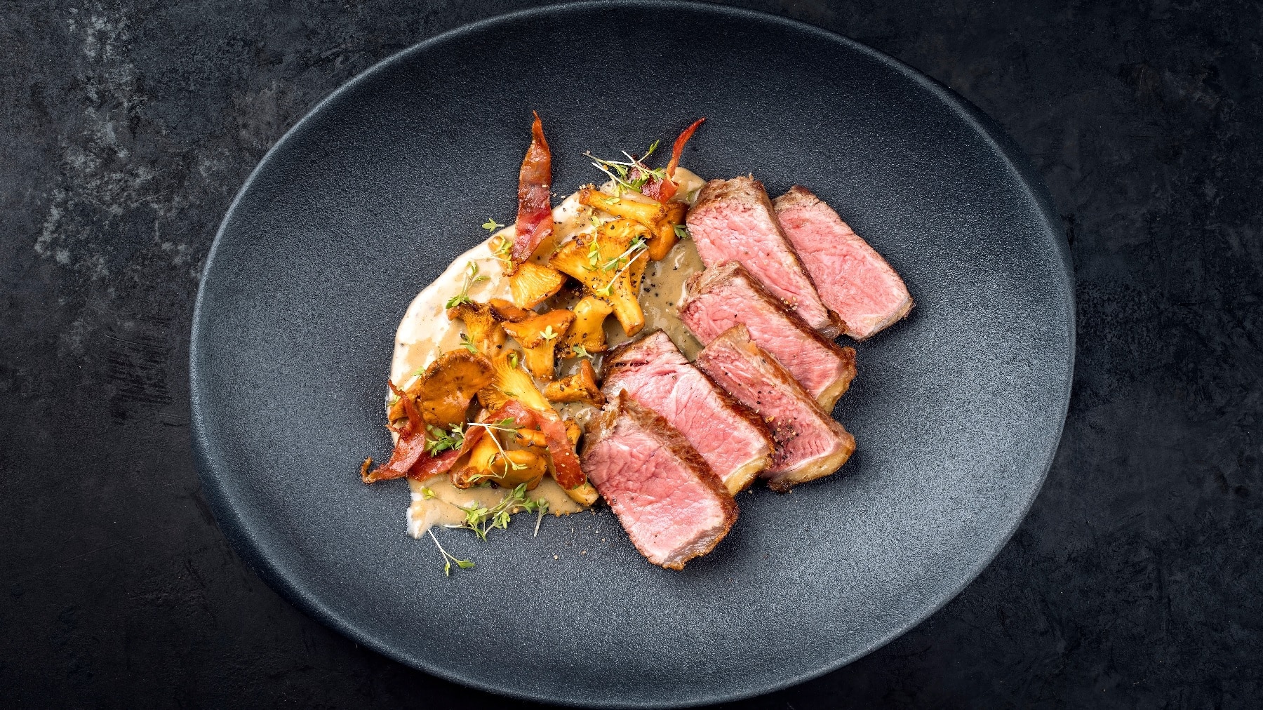 Ein in Scheiben geschnittenes Steak auf einem schwarzen Teller mit Sauce und den Pfifferlingen auf der linken Seite des Tellers.
