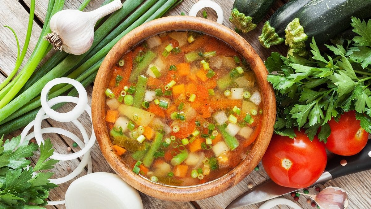 Aus der Vogelperspektive: Ein Topf grüne Bohnensuppe. Drumherum liegen Zucchini, Zwiebeln, Knoblauch und Tomaten.