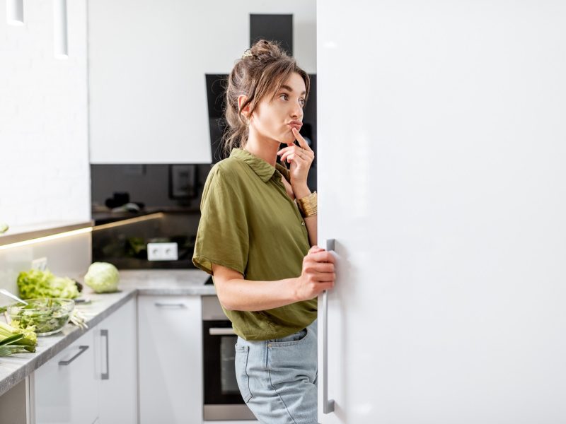 Eine Frau steht nachdenklich vor einem geöffneten Kühlschrank und überlegt, wie sie den Kühlschrank richtig einräumt.