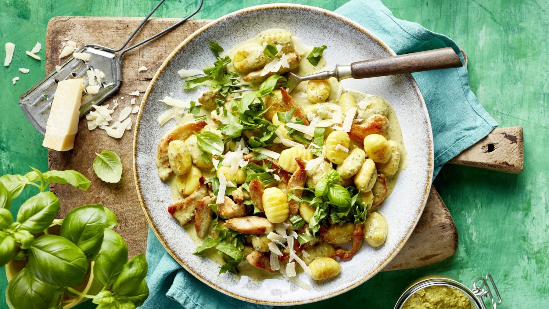 Pesto-Hähnchen-Geschnetzeltes mit Gnocchi auf einem Teller neben Parmesan und Basilikum auf einem Holzbrett vor grünem Hintergrund.