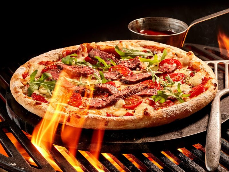 Pizza grillt auf Pizzastein und Kohlegrill. Daneben Pizzaschieber und Metallkelle. DRaufsicht.