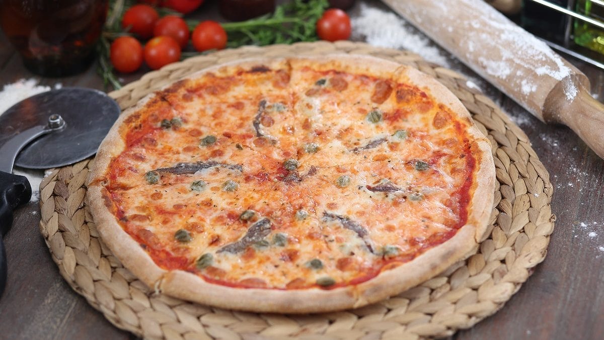 Draufsicht: eine Pizza mit Sardellen liegt auf einem Holzbrett. Grumherum liegen Tomaten und ein Nudelholz.