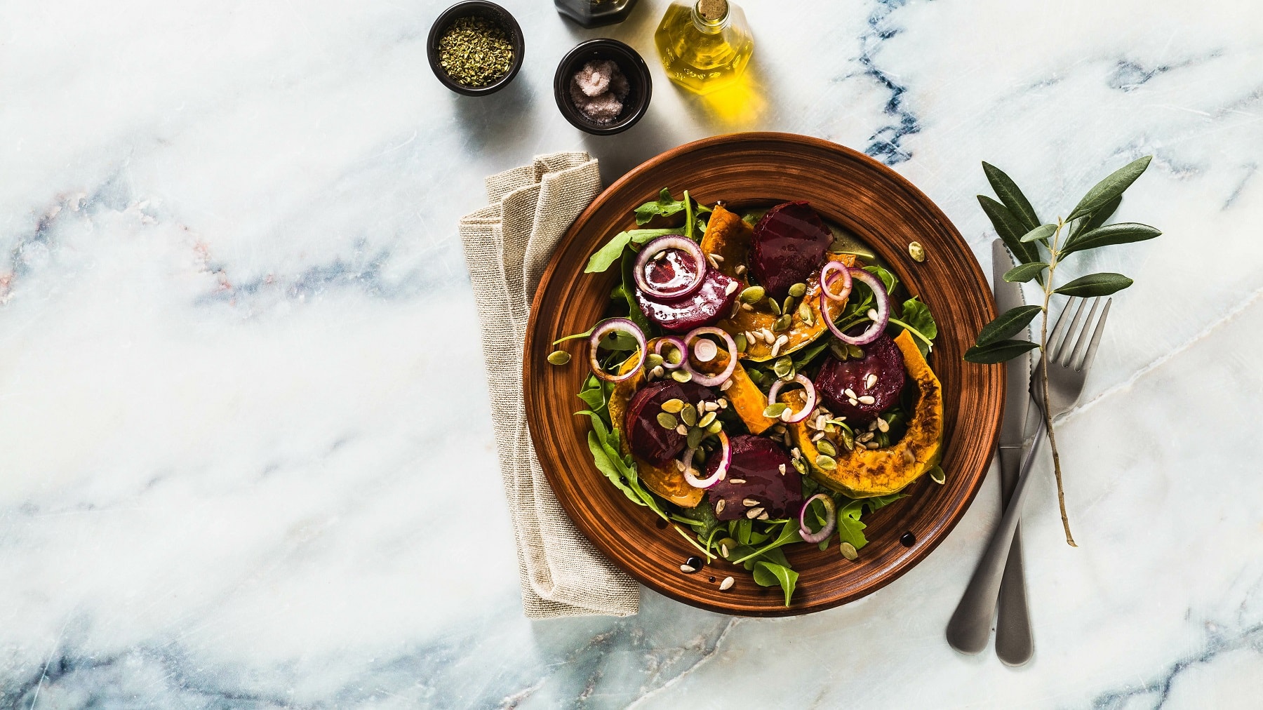 Ein Teller Salat mit Kürbis und Rote Bete aus dem Ofen in der Draufsicht, daneben liegen Messer und Gabel, außerdem eine Serviette und Ölflaschen.