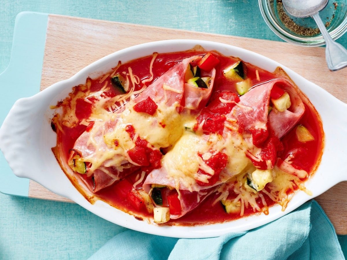 Mit Käse überbackene Schinken-Cannelloni, gefülllt mit Zucchini und serviert in einer defigen Tomatensauce in einer Auflaufform auf einem hellblauen Tisch.