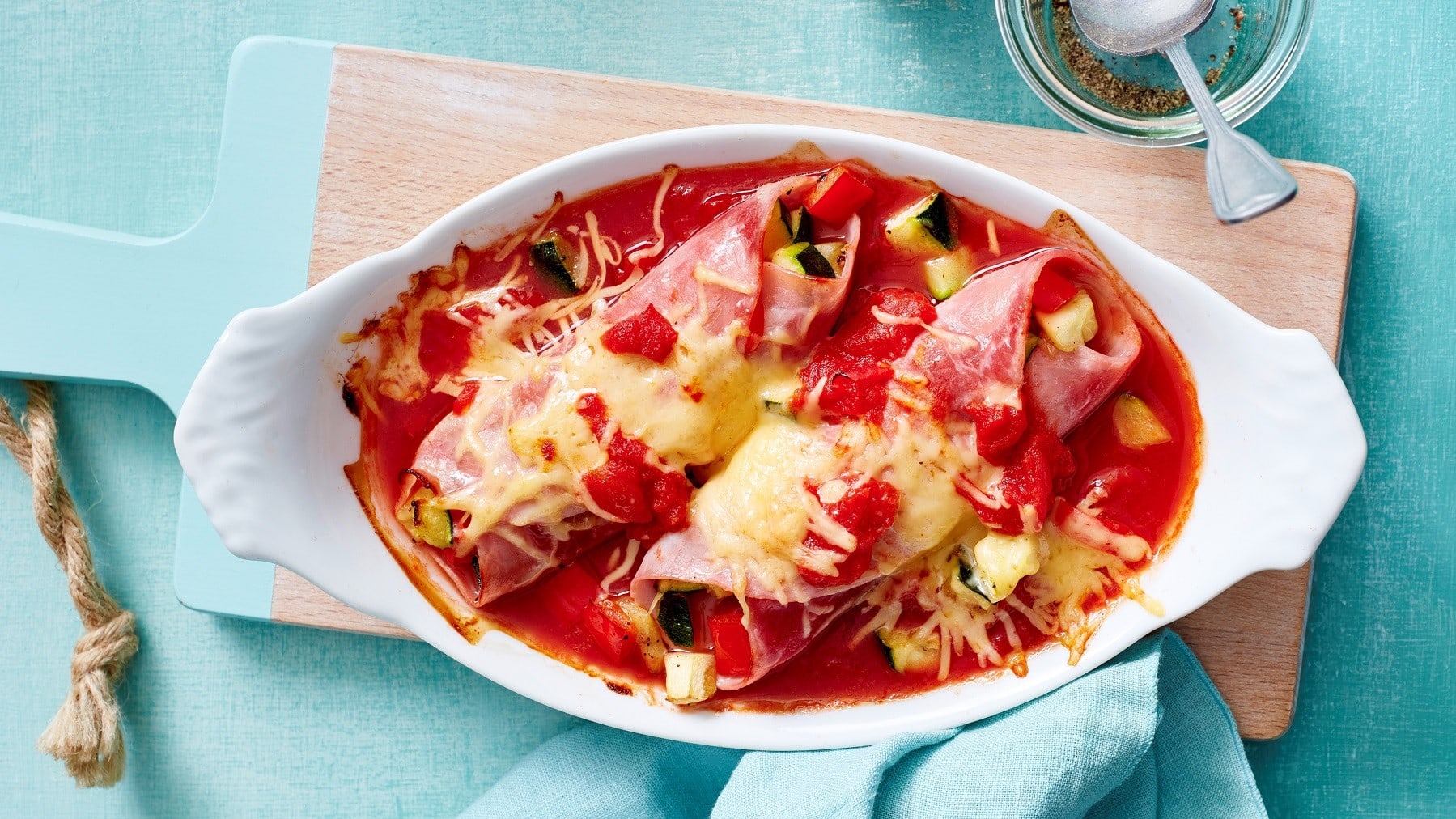 Mit Käse überbackene Schinken-Cannelloni, gefülllt mit Zucchini und serviert in einer defigen Tomatensauce in einer Auflaufform auf einem hellblauen Tisch.