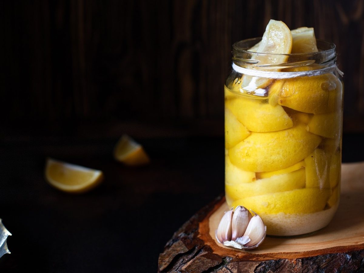Schnelle Salz-Zitronen, gelagert in einem Einmachglas mit Olivenöl und Knoblauch, geöffnet stehend auf einem Holzbrett vor dunklemn Hintergrund.